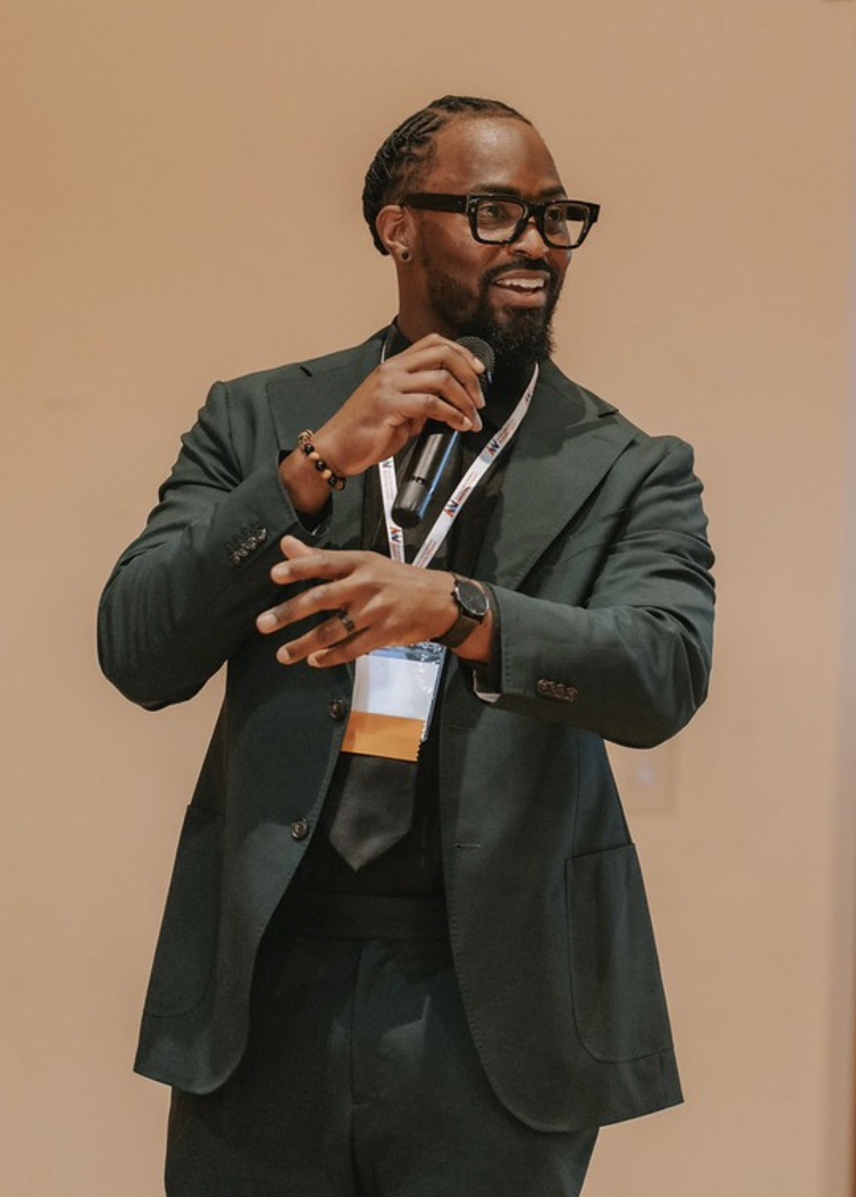 Man with glasses, beard, and braided hair smiling, wearing a dark blazer and lanyard, gesturing with hands.