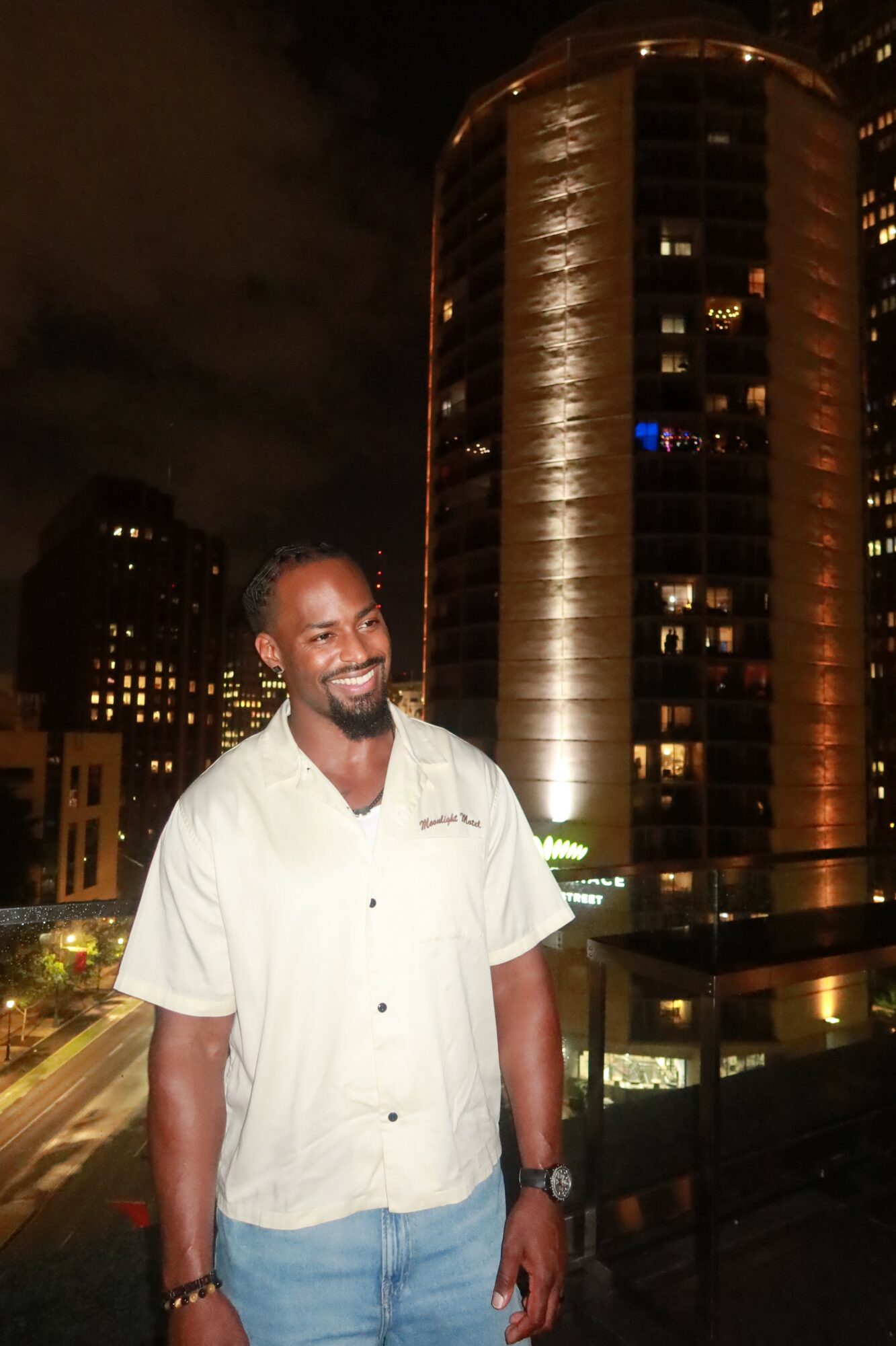 Smiling man in a white shirt and jeans standing on a balcony at night with tall illuminated buildings in the background.