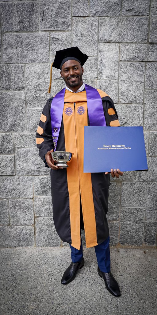 Person in graduation gown and cap holding a diploma and a silver bowl, standing against a stone wall.