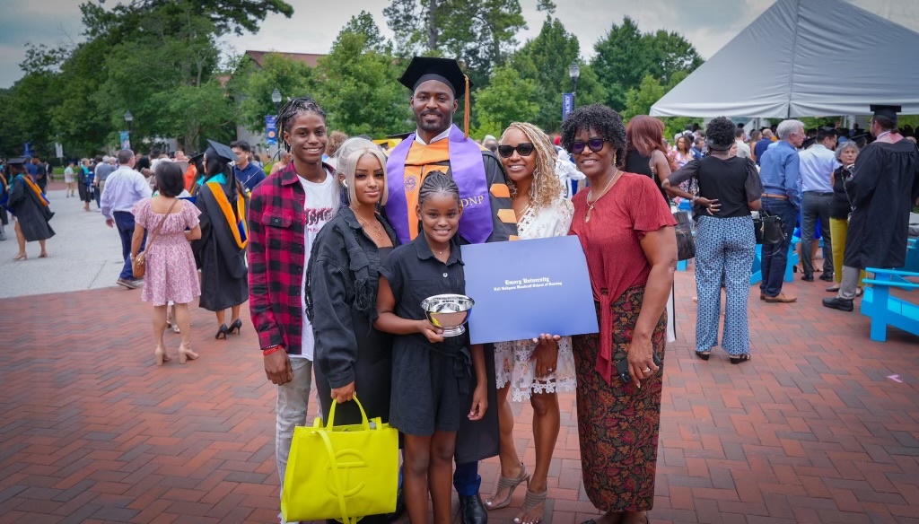 Group of six people, including a graduate in cap and gown, holding a diploma and a trophy, outdoors at a celebration.