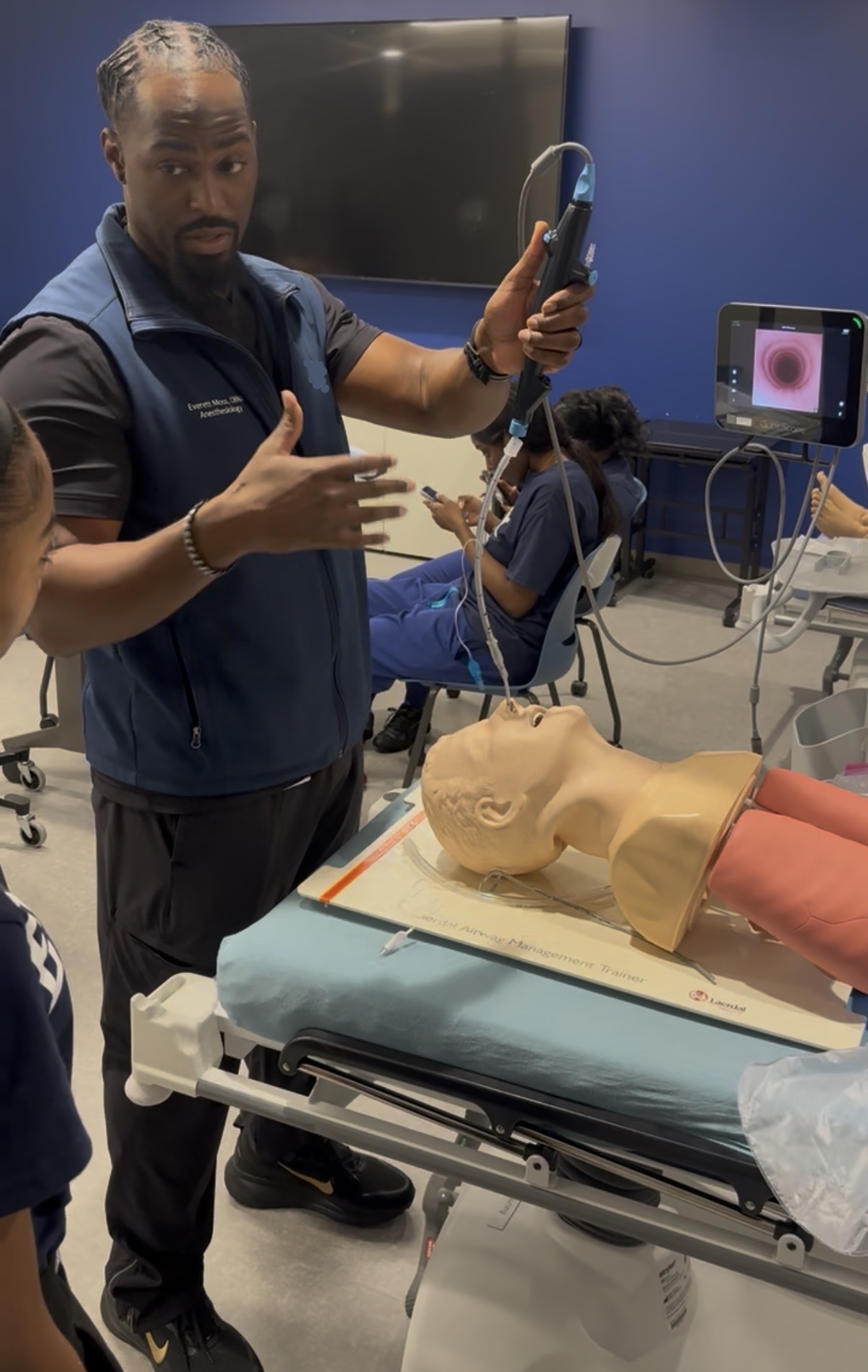 Instructor demonstrating medical procedure with a model arm and ultrasound equipment in a classroom setting.