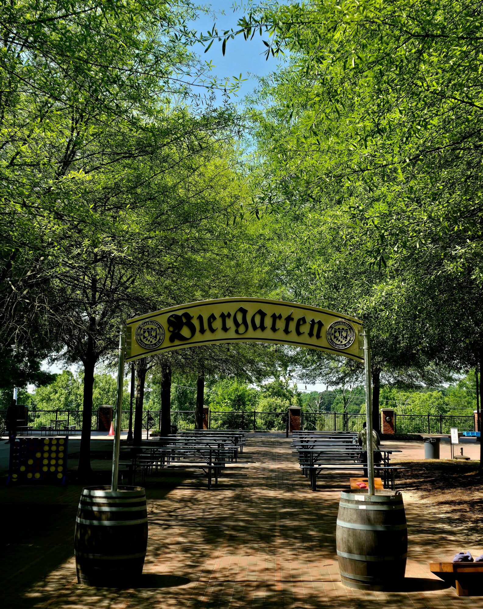 Entrance to a beer garden with a sign reading 'Biergarten' under trees, benches, and a clear sky.