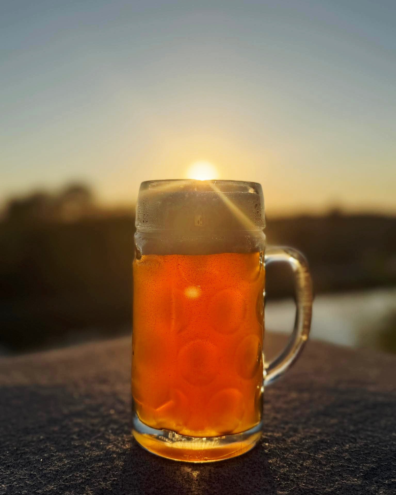 A glass mug of beer with foam on top, placed on a surface outdoors during sunset, with a blurred background.