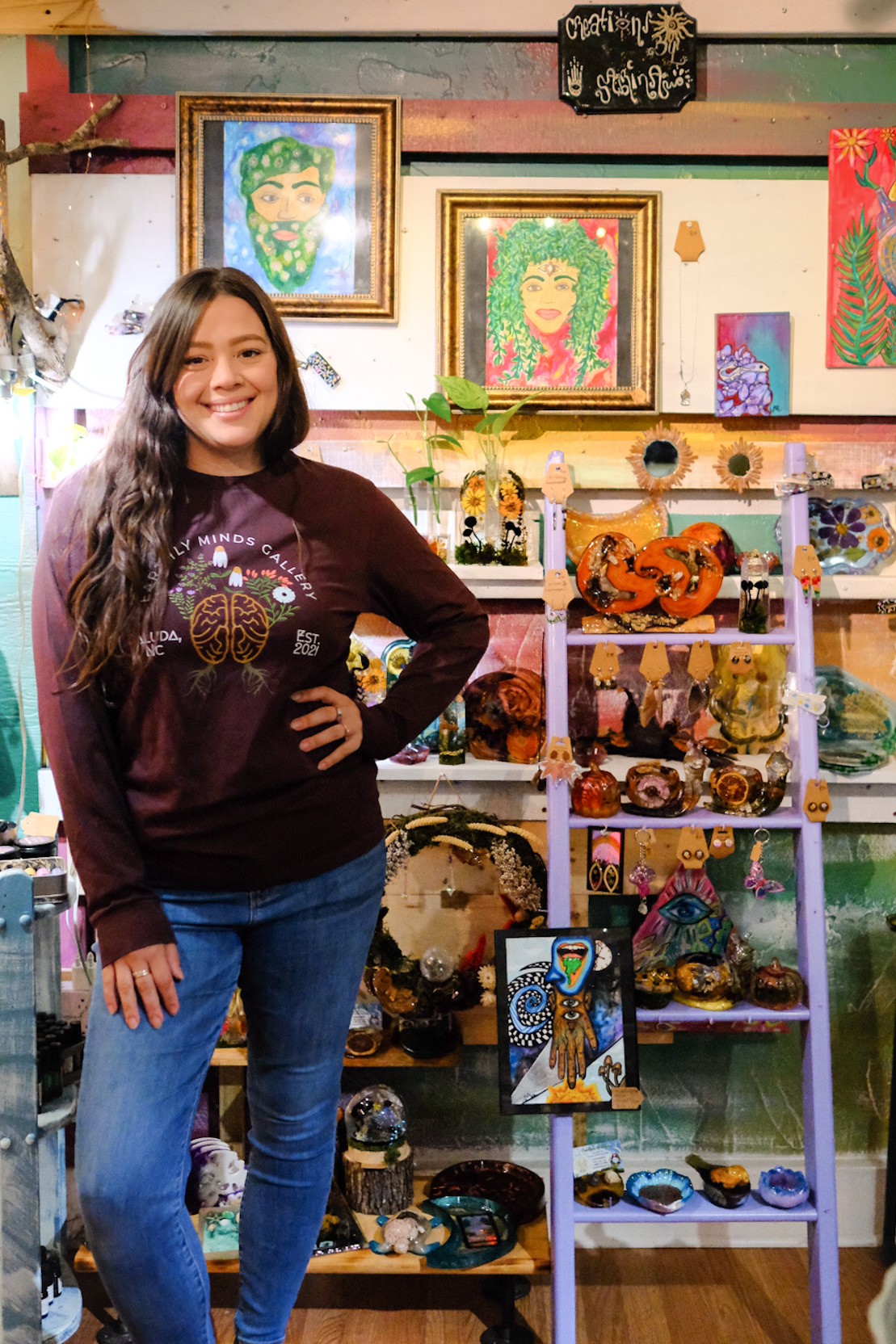 Young woman with long dark hair smiling in an art and craft store, surrounded by colorful paintings and handmade items.