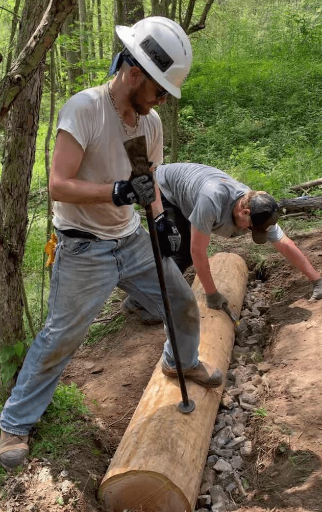Two people working outdoors on a log, one using a tool, surrounded by trees and greenery.