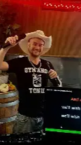Person wearing a cowboy hat and apron, smiling, standing indoors with a wooden ceiling and red lighting.