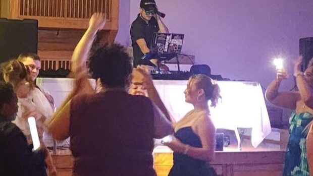 Group of people dancing and socializing at an indoor event with a DJ in the background.