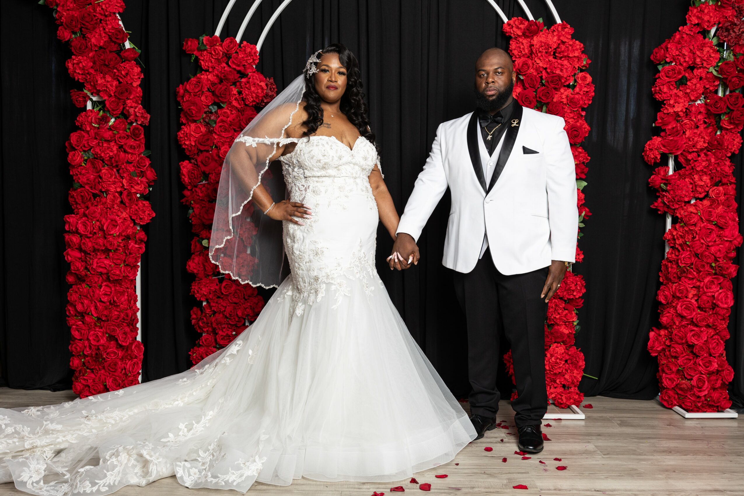 Bride and groom holding hands in front of red floral arrangements and black curtain backdrop, bride in white wedding gown, groom in white tuxedo jacket.