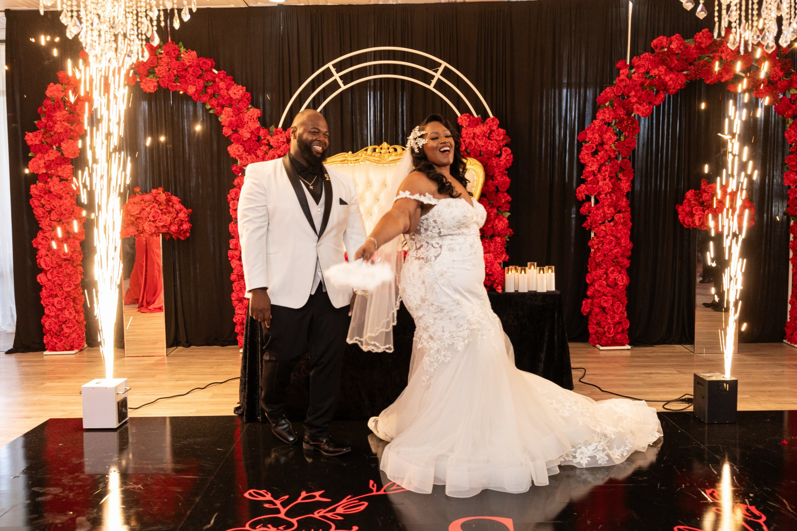 A bride and groom stand together in front of a decorated backdrop with red flowers and arches, smiling at the camera.