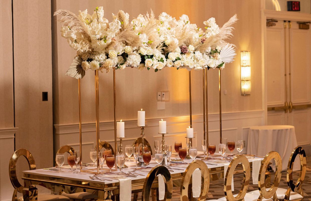 Elegant table setting with tall floral centerpiece, candles, and gold chairs in a decorated room.