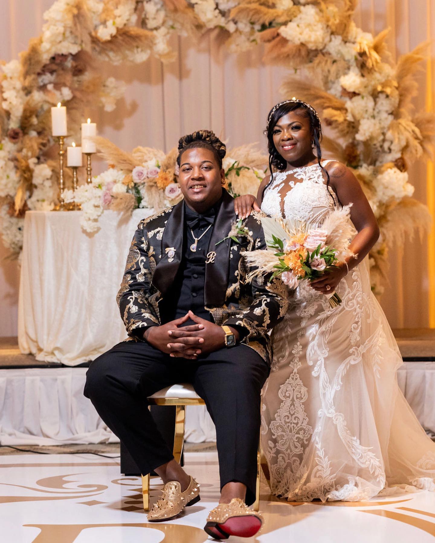 A couple at a wedding, the man seated and the woman standing, with floral decorations and candles in the background.