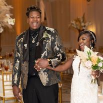 Two people smiling and holding hands at a decorated event, one in a floral blazer and the other in a white dress holding a bouquet.