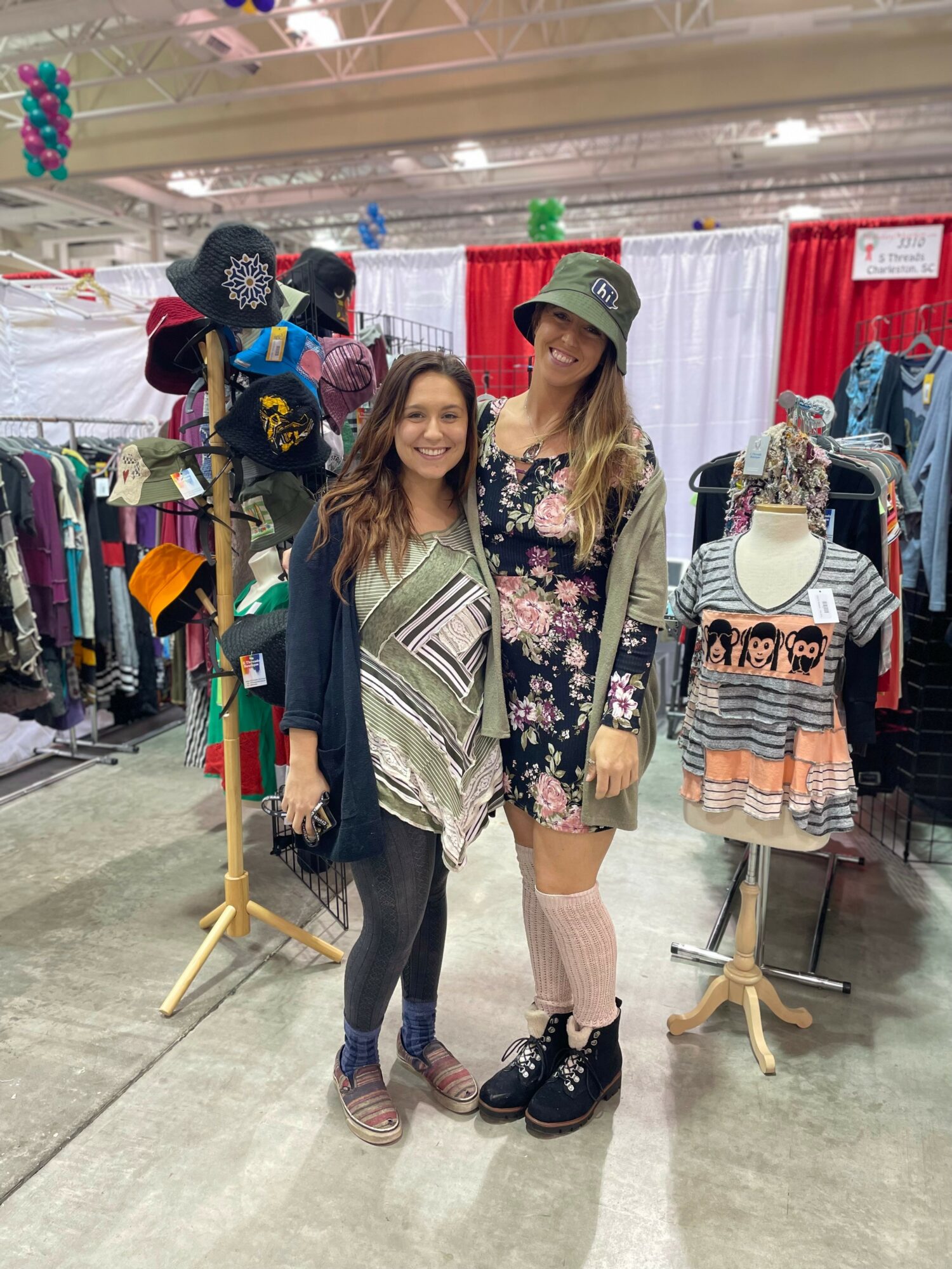 Two women stand together in a clothing store, smiling, surrounded by racks of clothes and accessories.