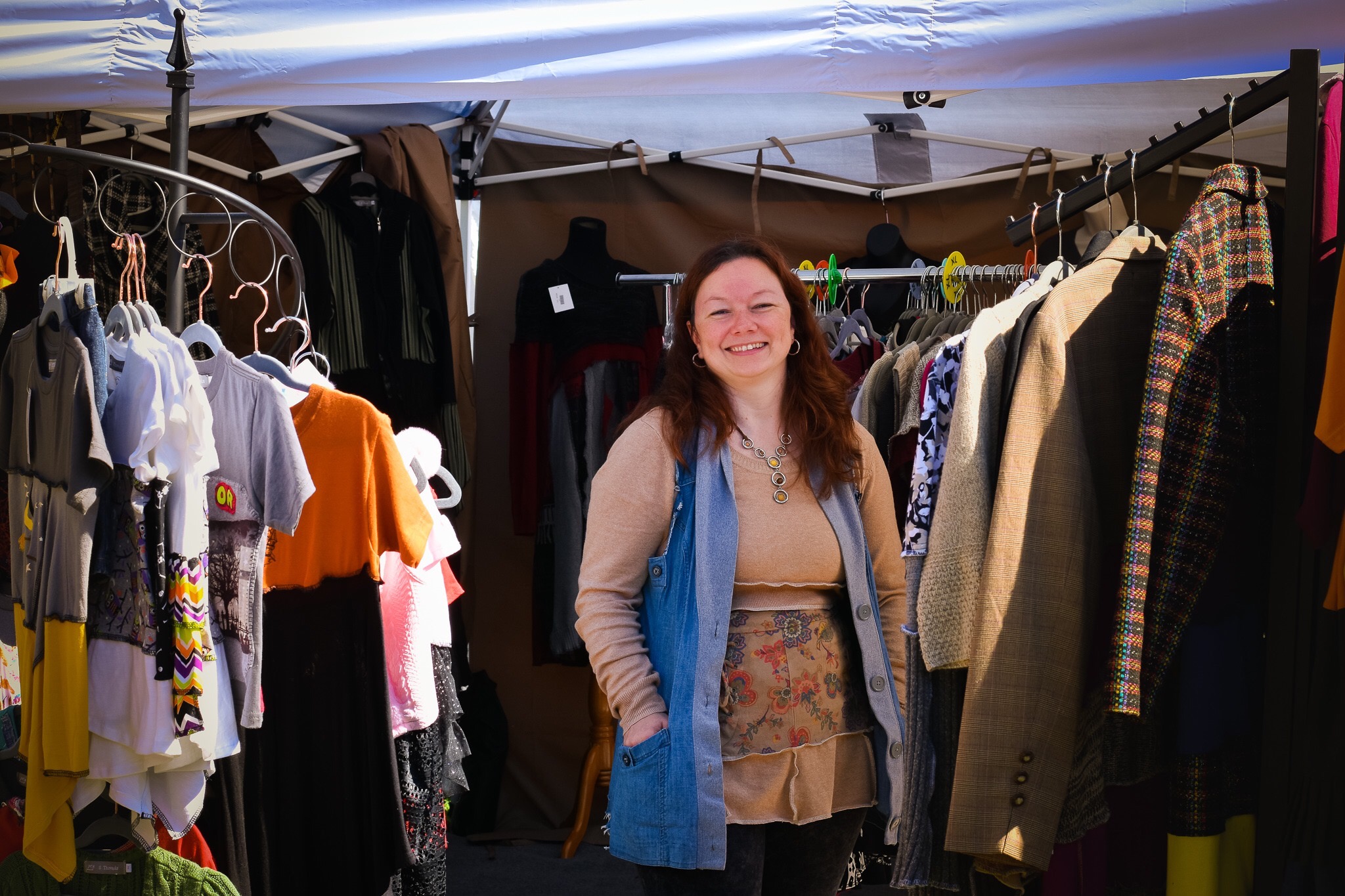 Woman smiling standing among clothing racks at an outdoor market, wearing a beige top and blue vest.