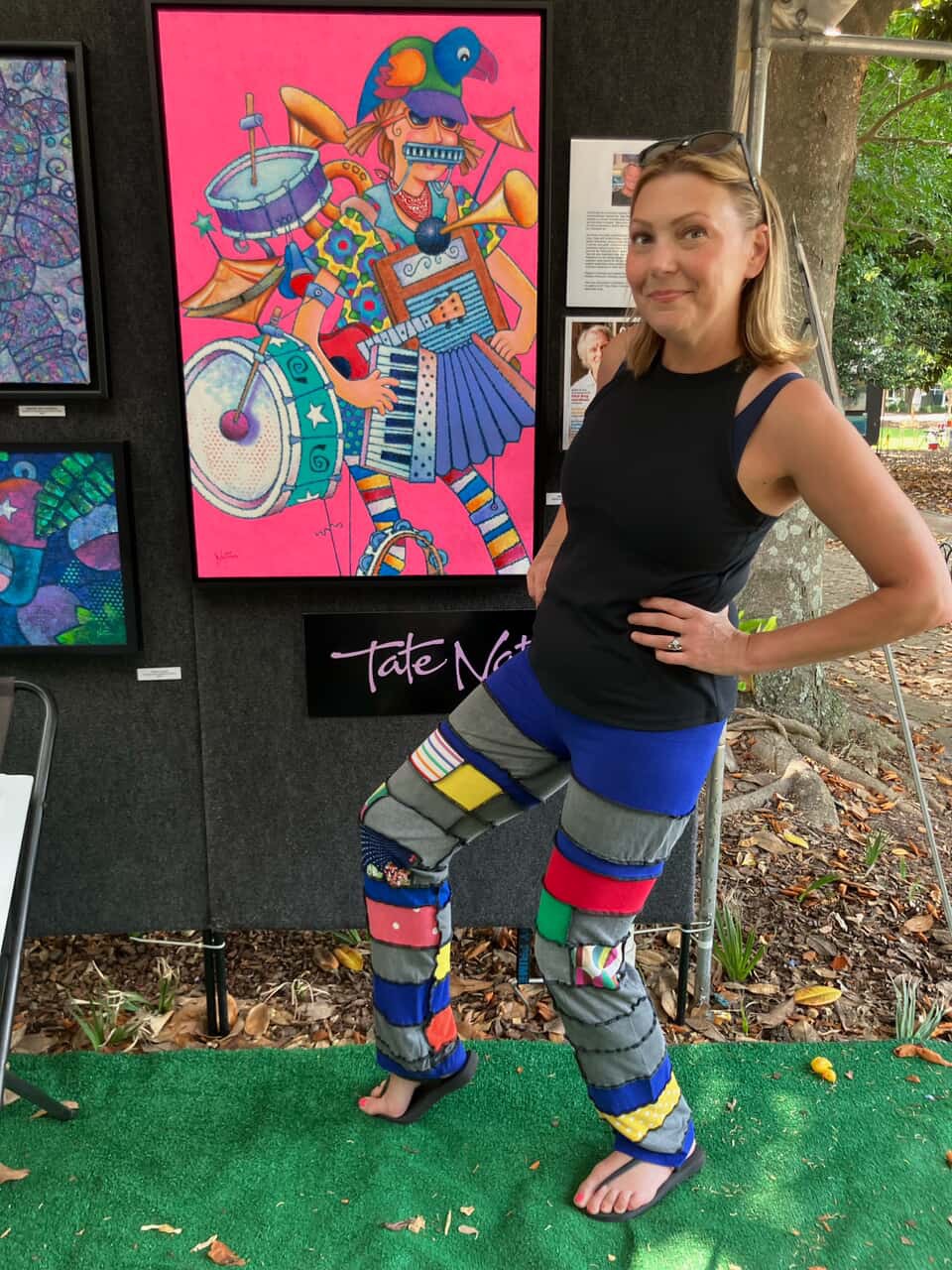 Young woman with long hair in a black tank top and colorful patchwork pants standing outdoors, smiling, with artwork behind her.