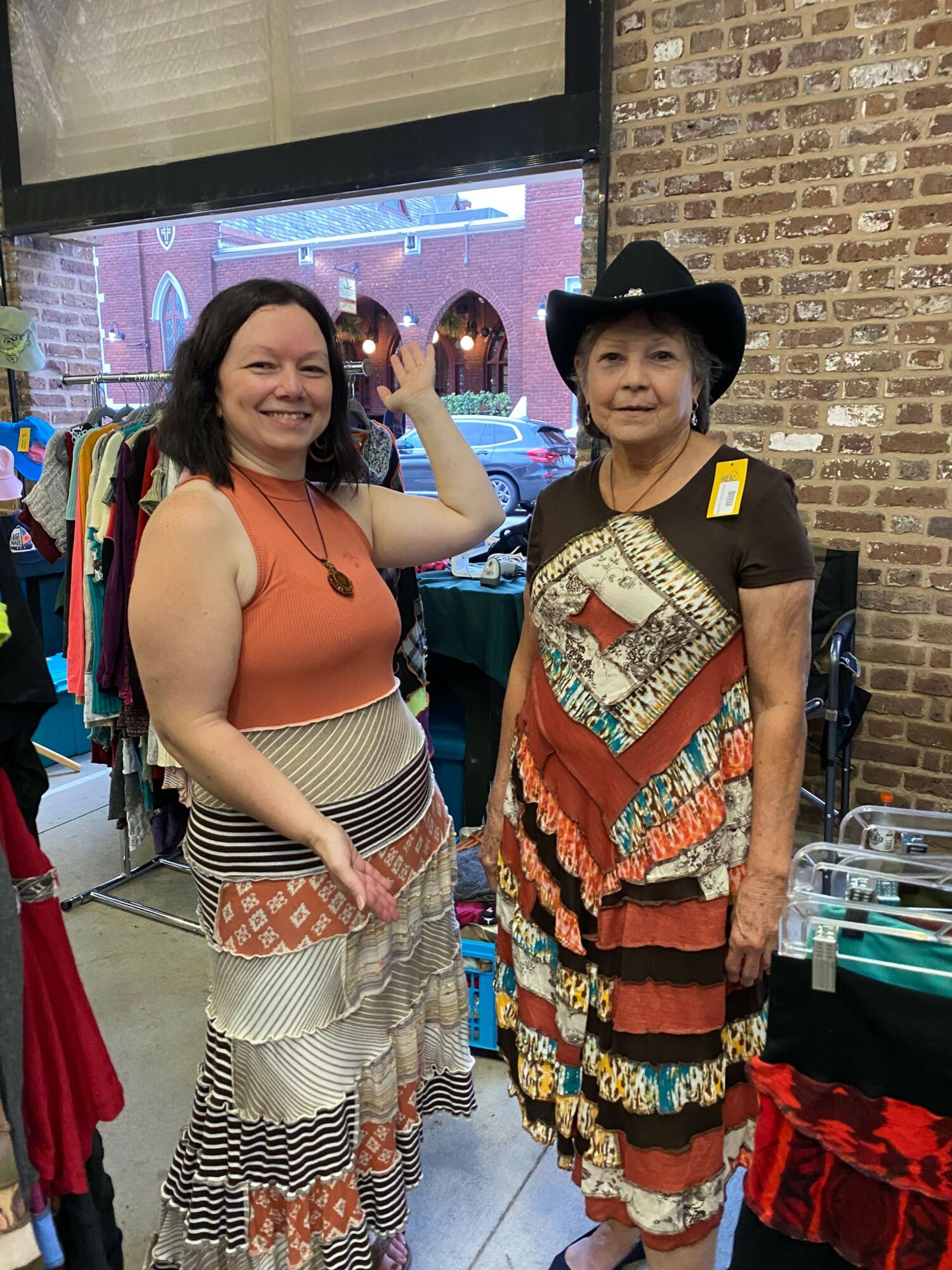 Two women standing indoors, one wearing a black hat and colorful dress, the other in a sleeveless top and patterned pants.