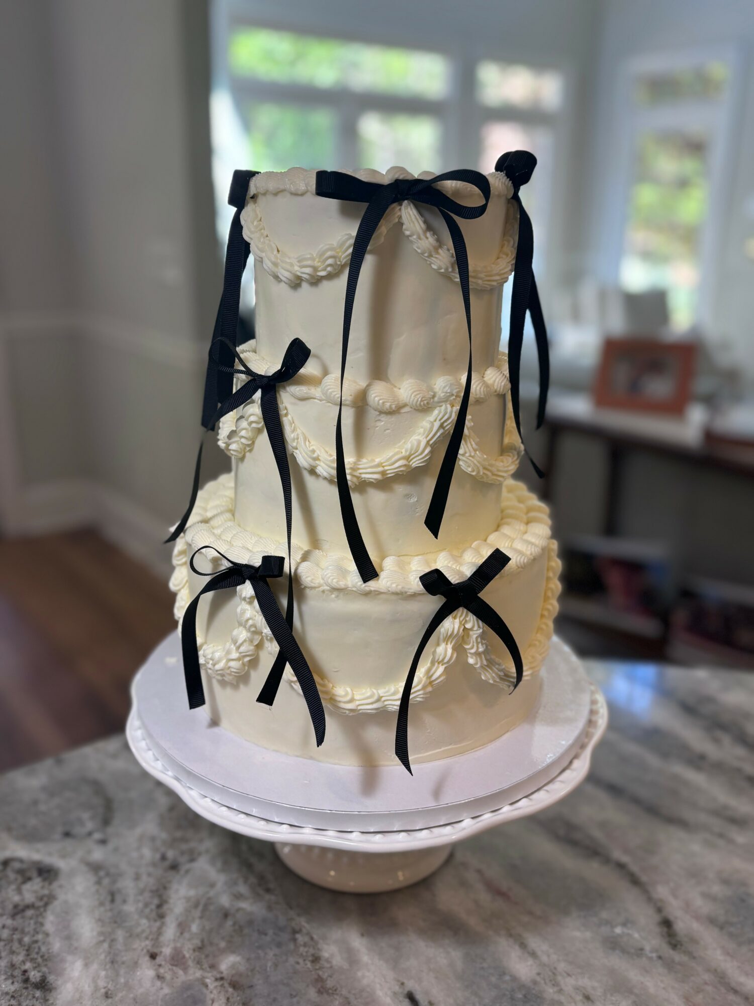 Three-tiered cake with white frosting and black ribbon bows, displayed on a white cake stand in a room with large windows.