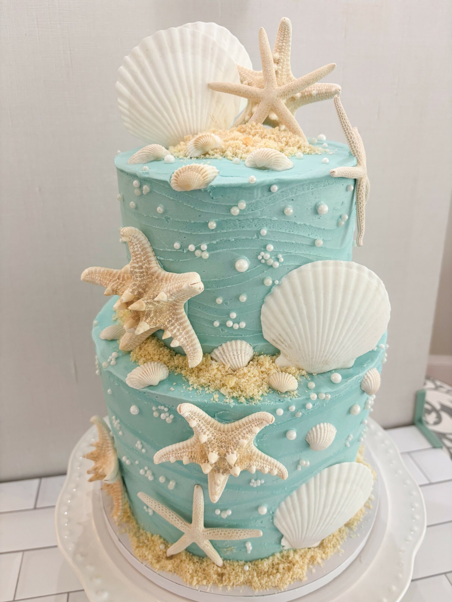 Two-tiered cake decorated with seashells, starfish, and pearls, featuring a light blue ocean theme, on a white cake stand.