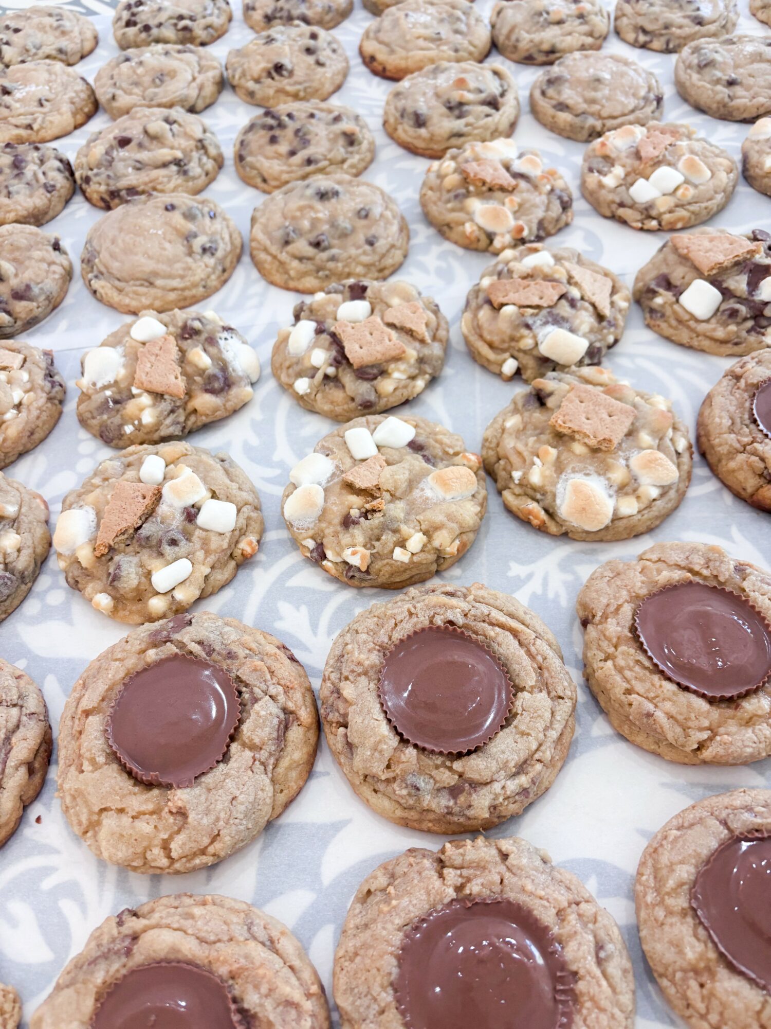 Rows of cookies with chocolate centers and toppings on a patterned surface, some with white sugar crystals, some plain.