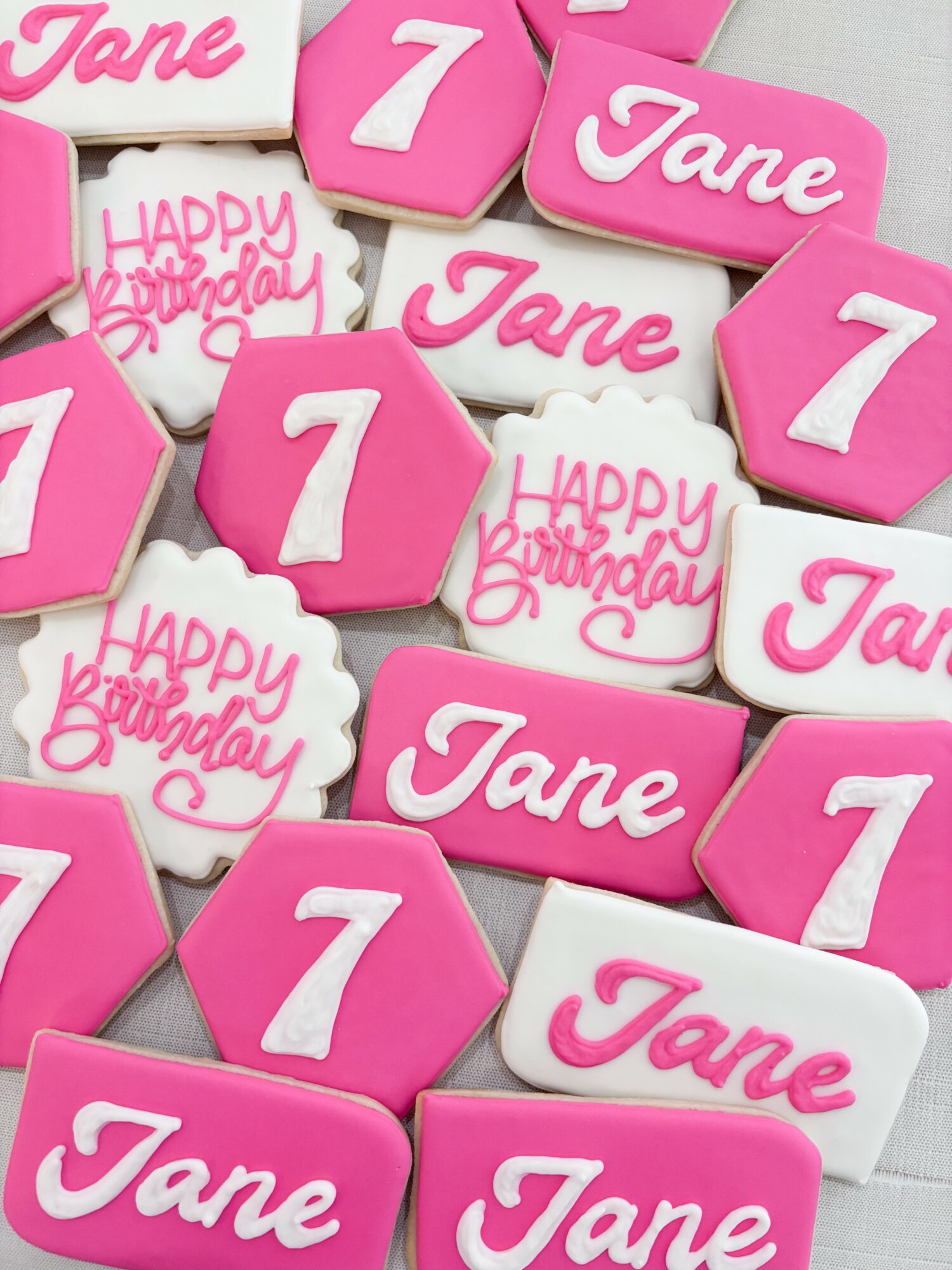 Decorated cookies with pink, white, and purple icing, featuring names, numbers, and birthday messages, arranged on a white surface.