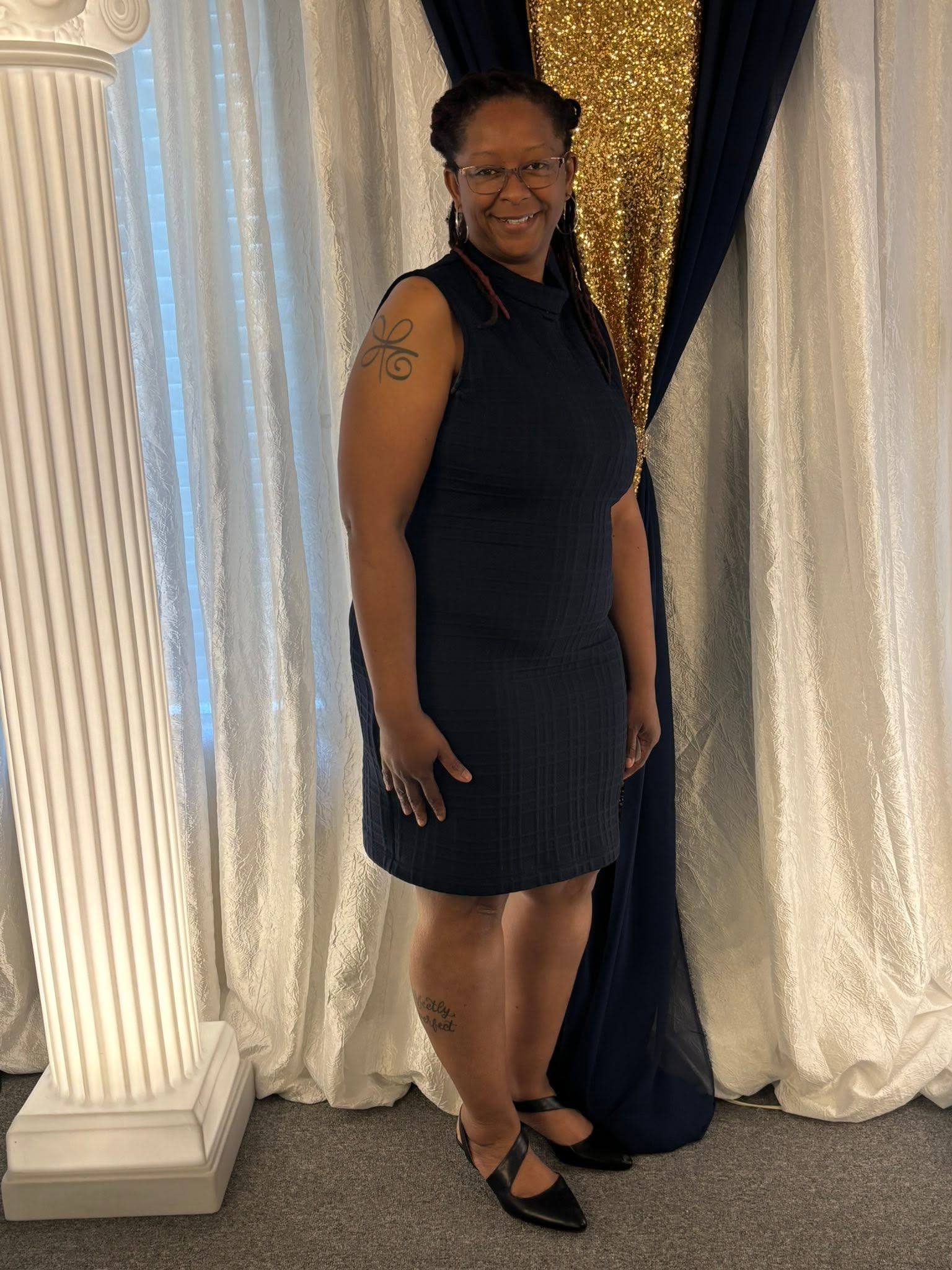 Woman in black dress standing in front of curtains, smiling, with glasses and tattoos on arms, wearing black heels.