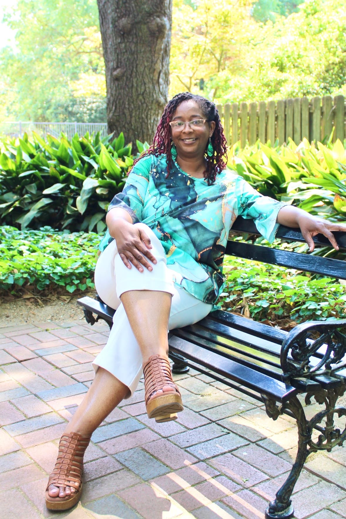 Woman sitting on a park bench, smiling, with greenery and a tree in the background.