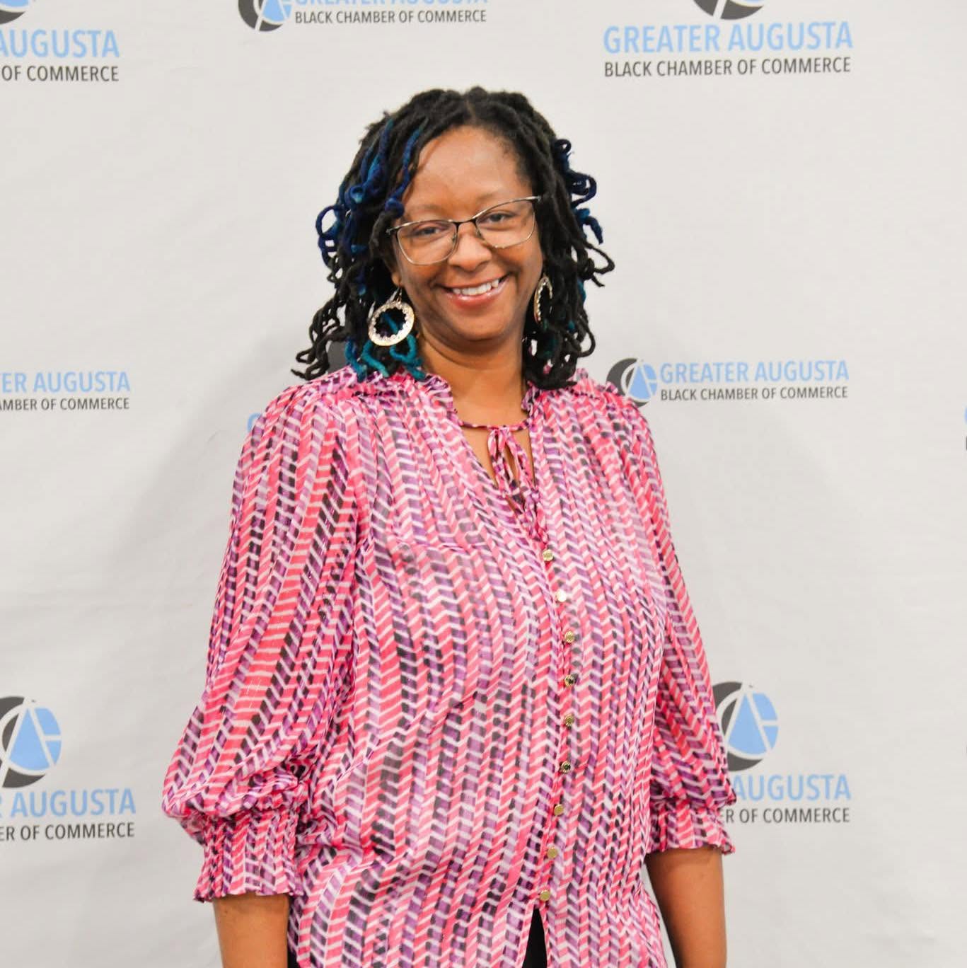Smiling woman with glasses and earrings, wearing a pink patterned blouse, standing in front of a backdrop with logos.