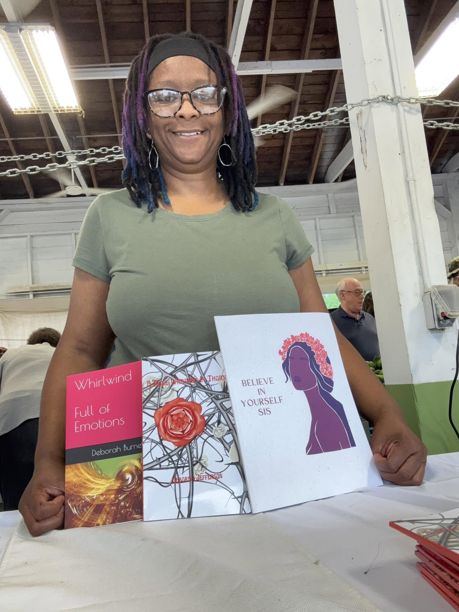 Smiling woman with glasses and purple-dyed hair holding two books, standing indoors with industrial ceiling and chain in background.