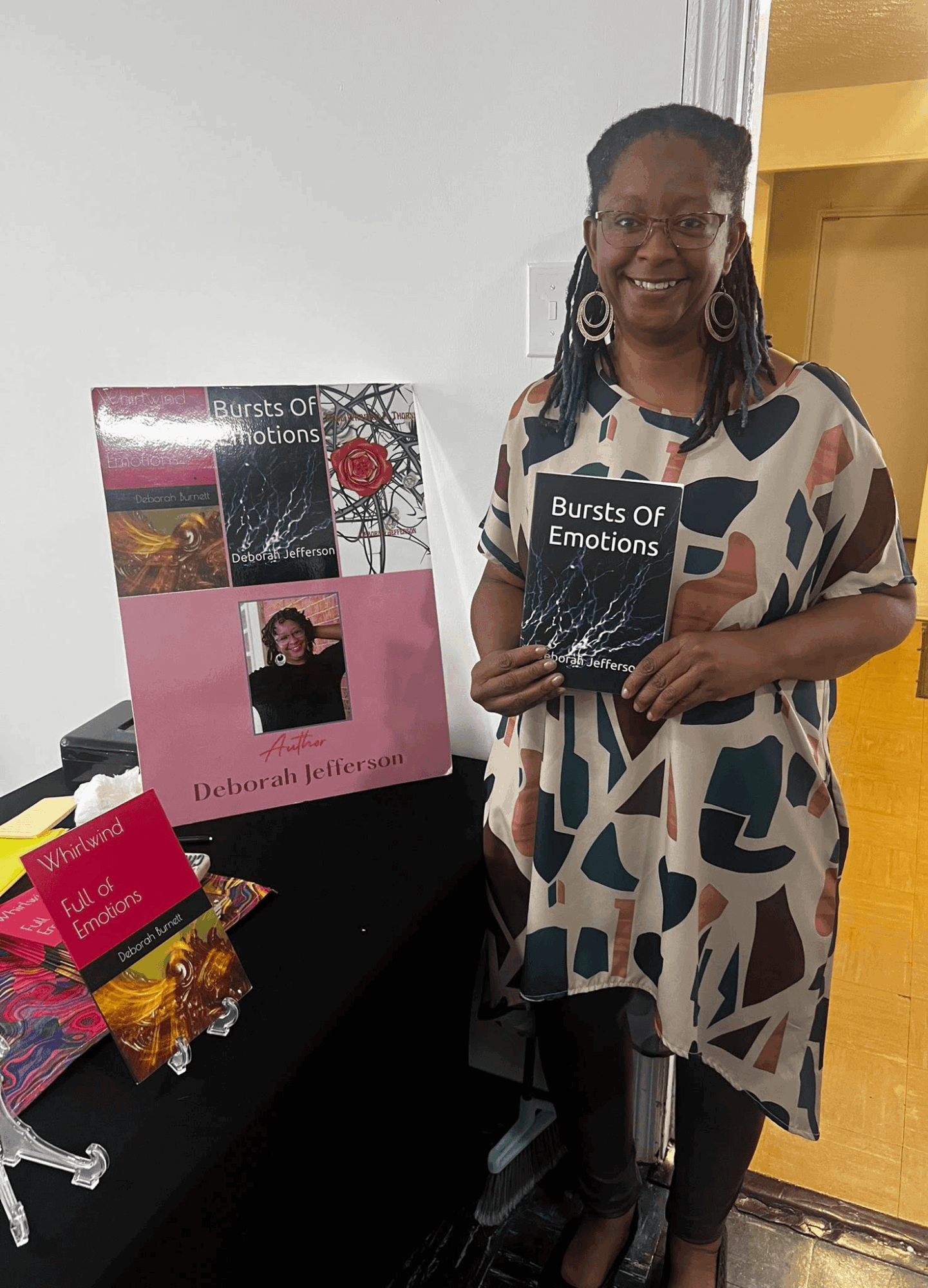 Woman holding a book titled 'Bursts of Emotions' standing next to a table with more books and a gift bag, smiling.