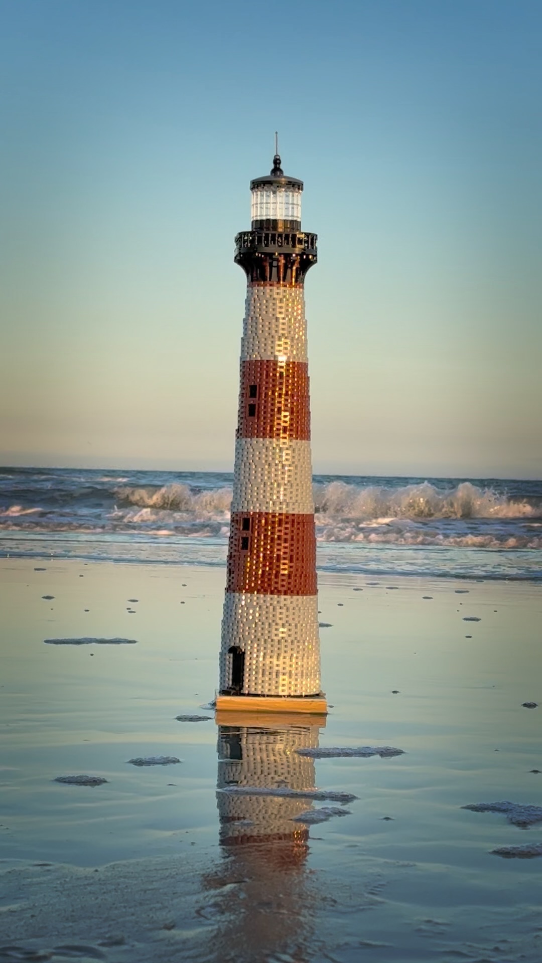 Lighthouse standing in shallow water with waves, sky in background, and reflection on wet sand.