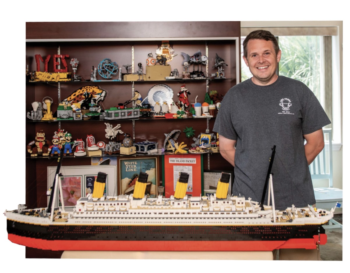 Man standing behind a large ship model with LEGO creations on shelves in the background.