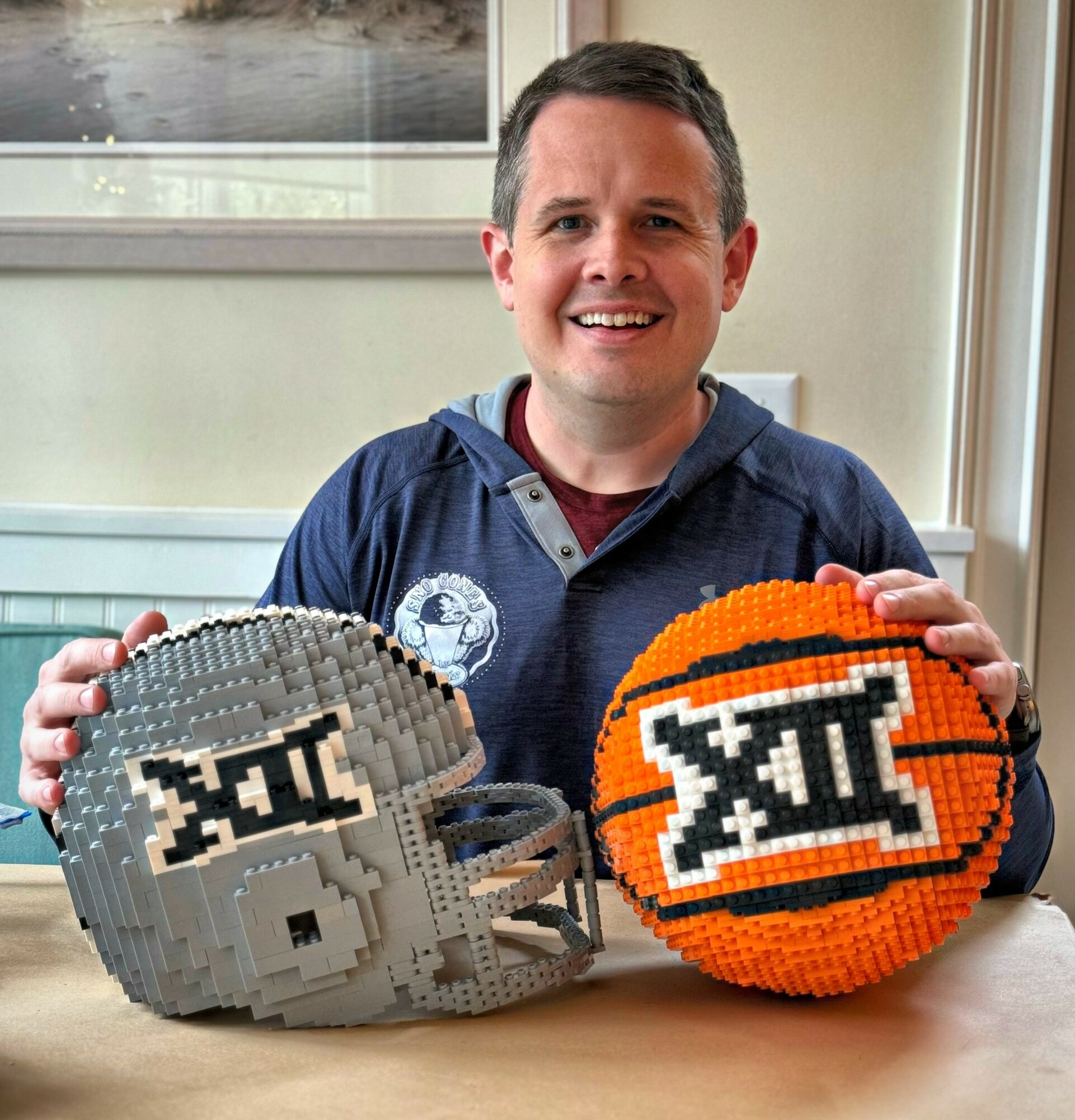 Smiling man holding a pixelated football and basketball with Big 12 logos, sitting at a table near a window.