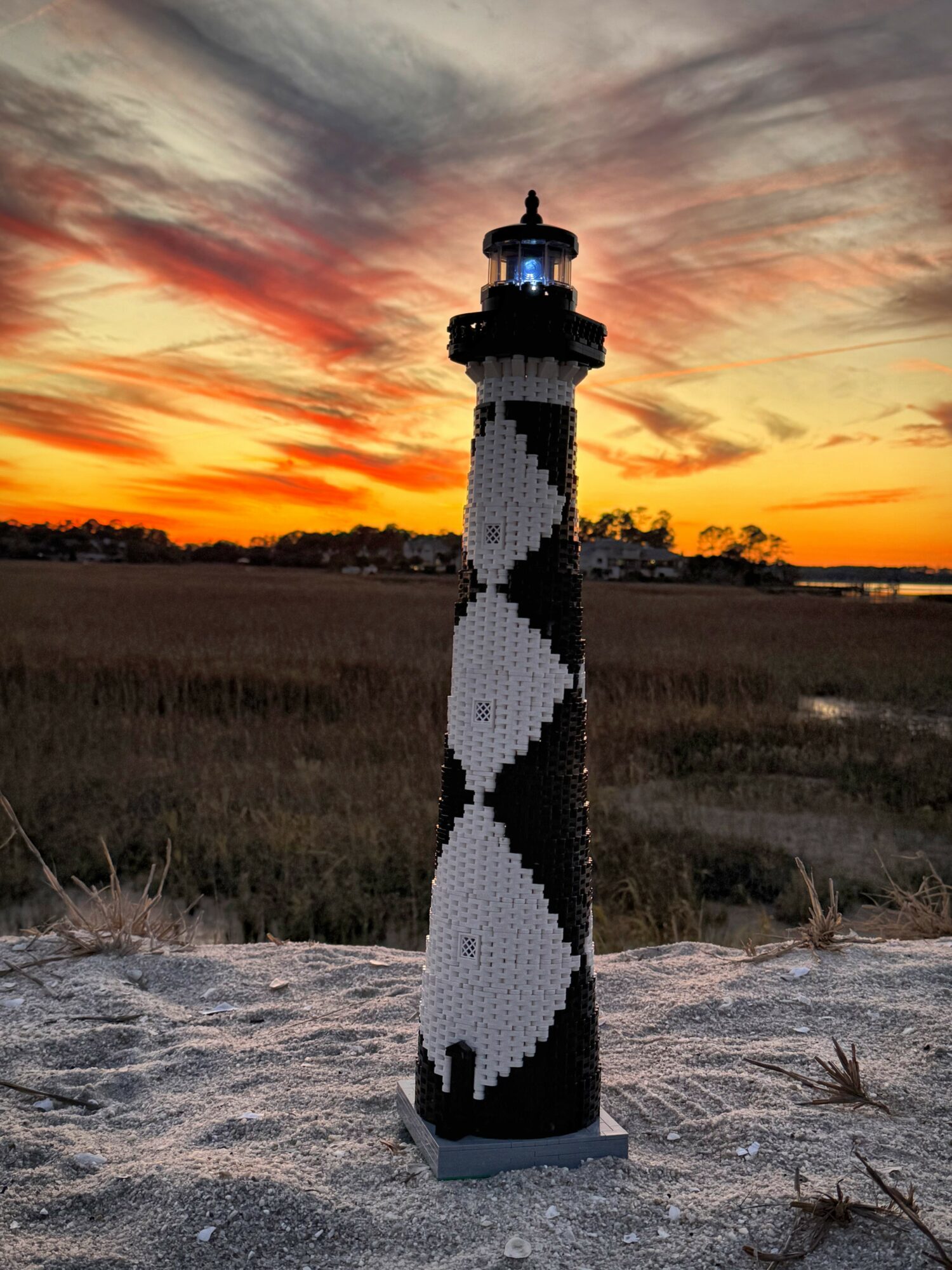 Lighthouse with black and white spiral pattern stands on sandy ground during sunset sky with orange and purple clouds.