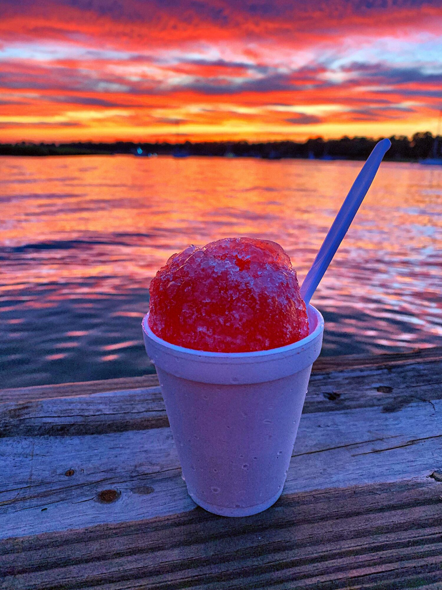 A cup of red shaved ice with a spoon on a wooden surface, sunset sky with colorful clouds and water in background.