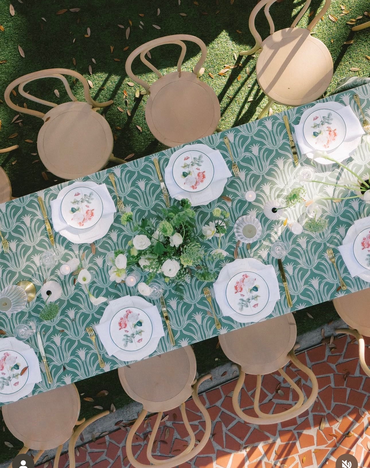 Table with a patterned tablecloth, white plates with floral designs, and beige chairs, outdoors on a brick surface.