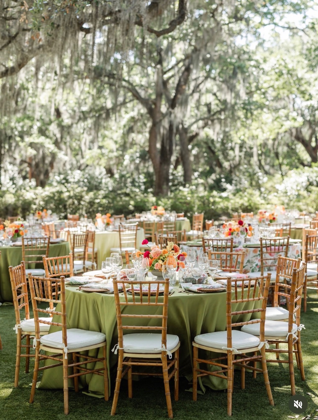 Outdoor event with round tables covered in green tablecloths, surrounded by wooden chairs, set with plates and floral centerpieces, under trees.