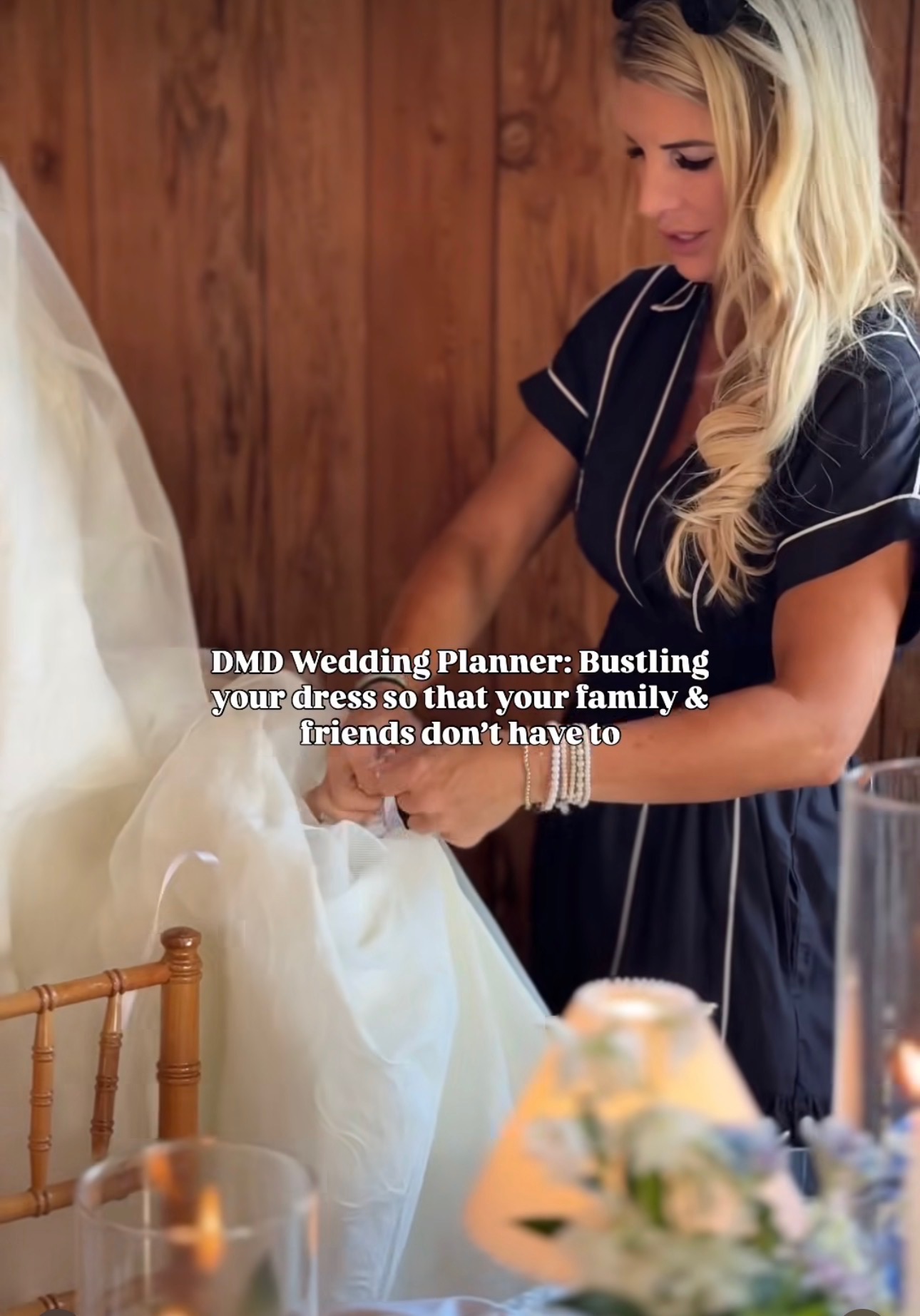 Woman with long blonde hair in a dark dress adjusts a white wedding dress on a mannequin, wooden background, flowers nearby.