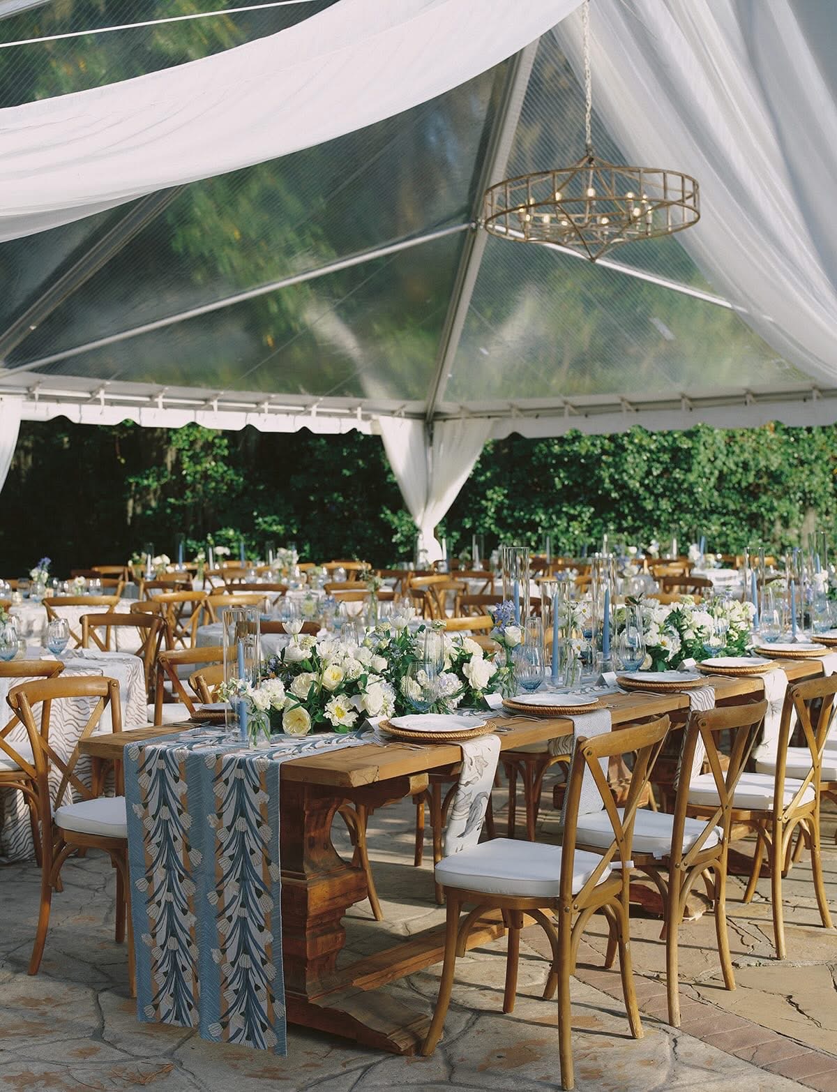 Wedding reception setup with long tables, chairs, floral centerpieces, under a white tent with greenery outside.