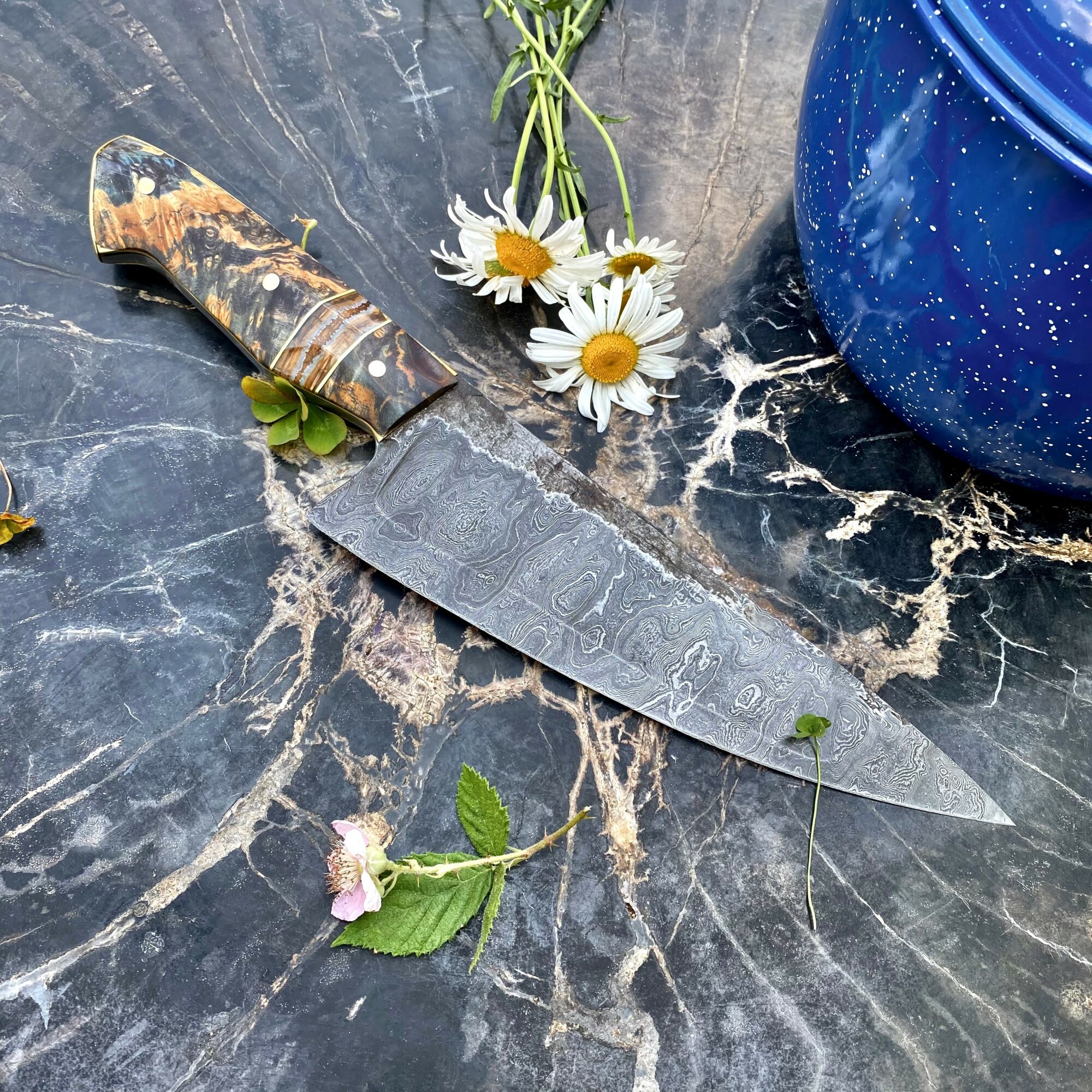 Knife with a patterned handle and a metal blade, placed on a dark marble surface with flowers and a blue pot nearby.