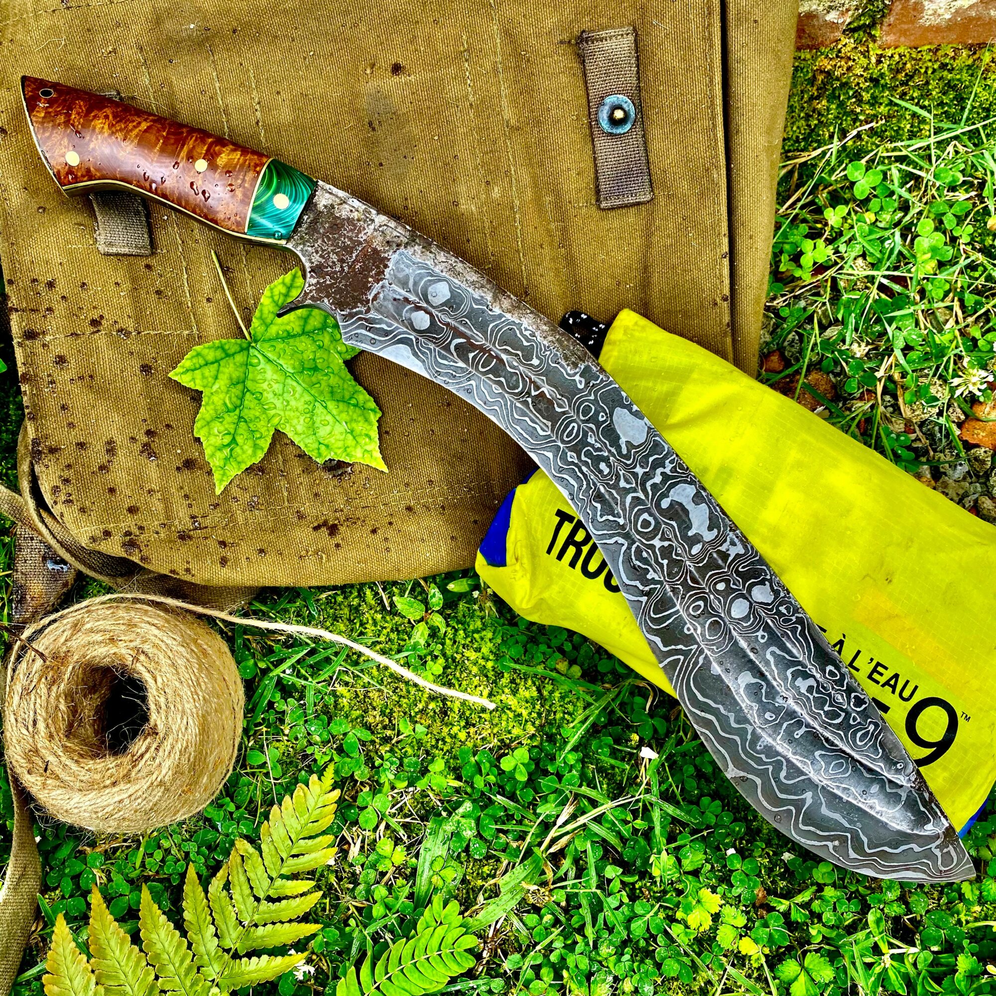Damascus-style patterned knife with a wooden handle, placed on a green outdoor surface with a yellow bag and plant leaves.