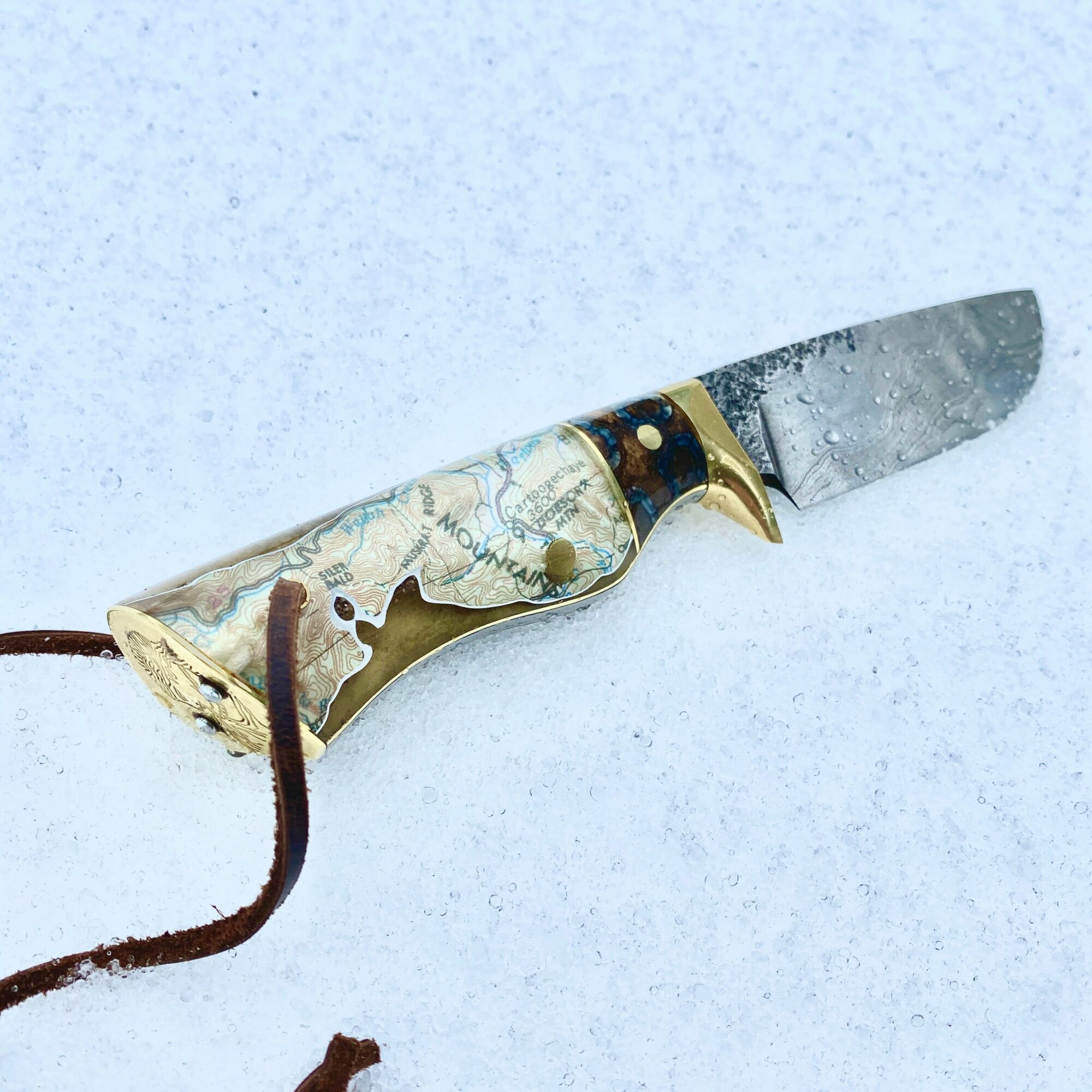A reflective knife with a gold handle lies on snow, with a brown leather strap attached. Alt_text word count: 20.