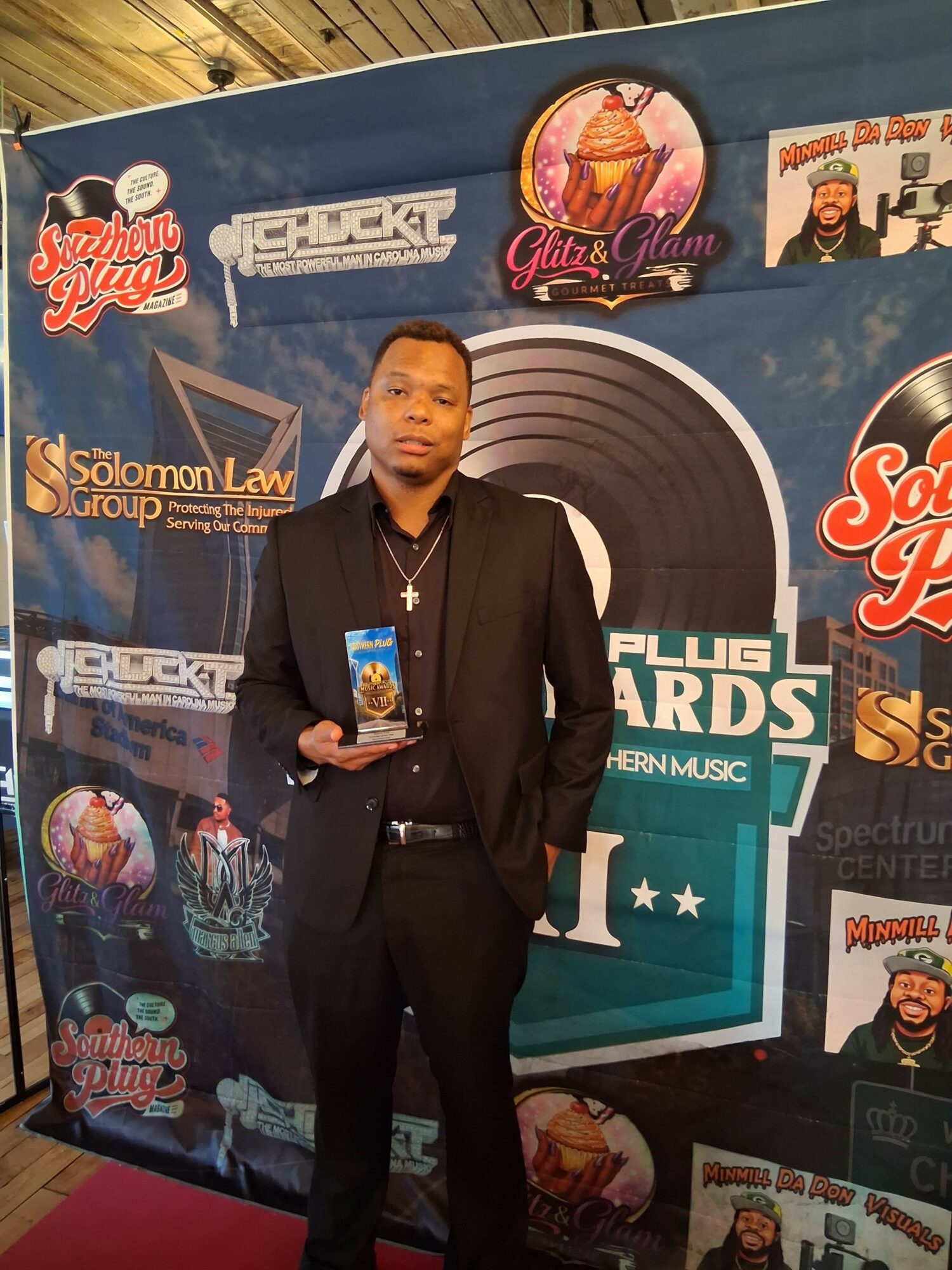 Man in black suit holding an award, standing in front of a colorful backdrop with logos and text.