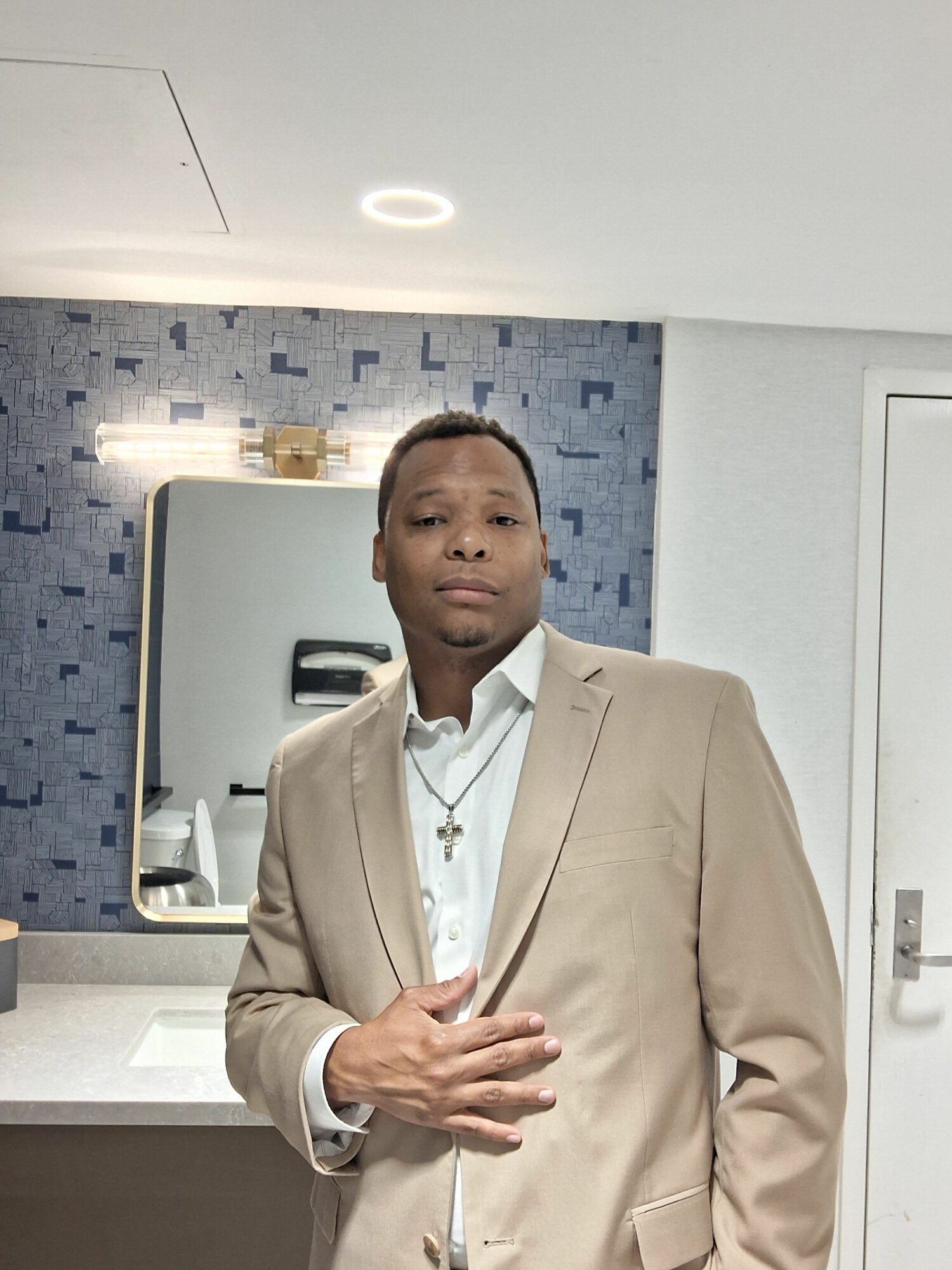 Man in beige suit standing indoors with a mirror and blue textured wall behind him.