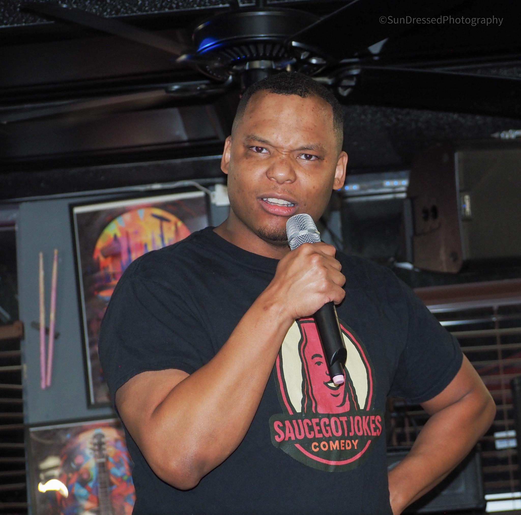 Man holding microphone, wearing black T-shirt with a graphic and text, standing indoors with a screen in background.