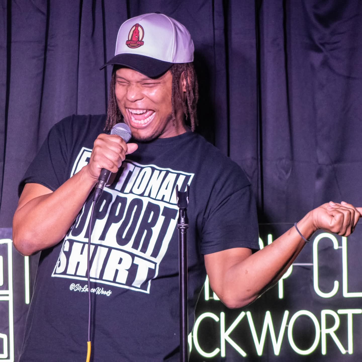Person laughing, holding microphone, wearing a black T-shirt and a white cap with a red logo, standing in front of a dark curtain.