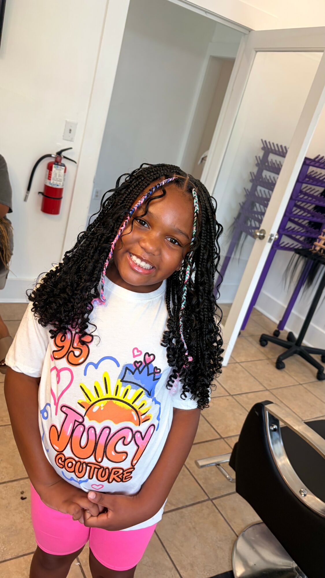 Smiling young girl with curly hair wearing a white T-shirt and pink shorts, standing indoors near a fire extinguisher.