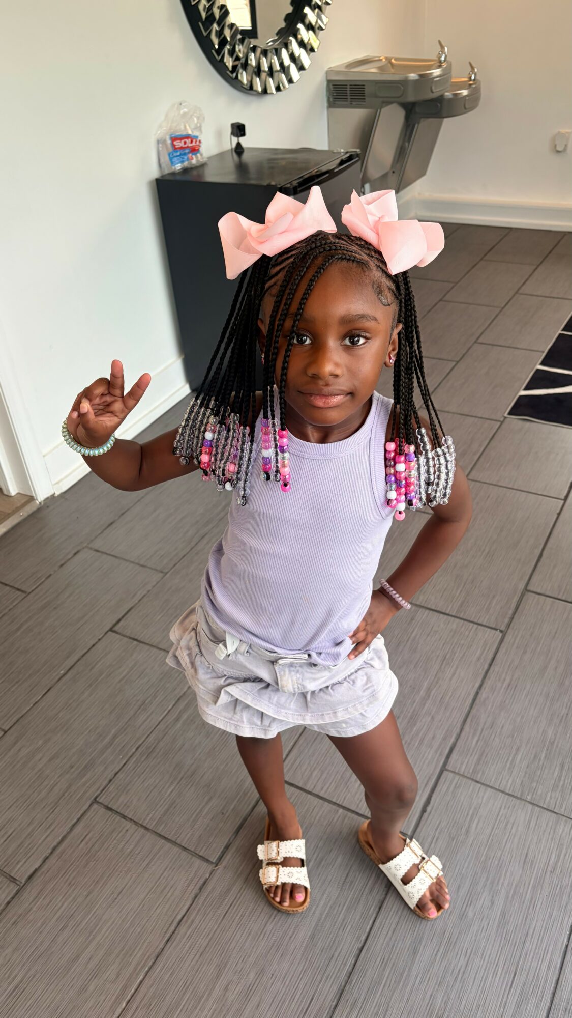 Young girl with braided hair and pink bows, standing indoors with one hand on her hip and the other raised, smiling.