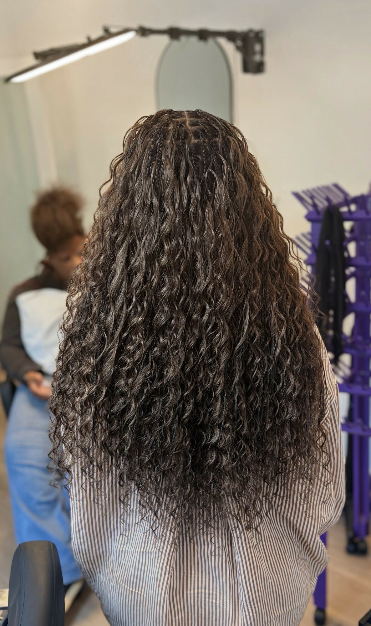 Back view of a person with long, curly hair in a salon, with a mirror and salon equipment visible.