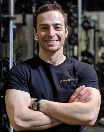 Man smiling with arms crossed in a gym, wearing a black t-shirt and smartwatch.