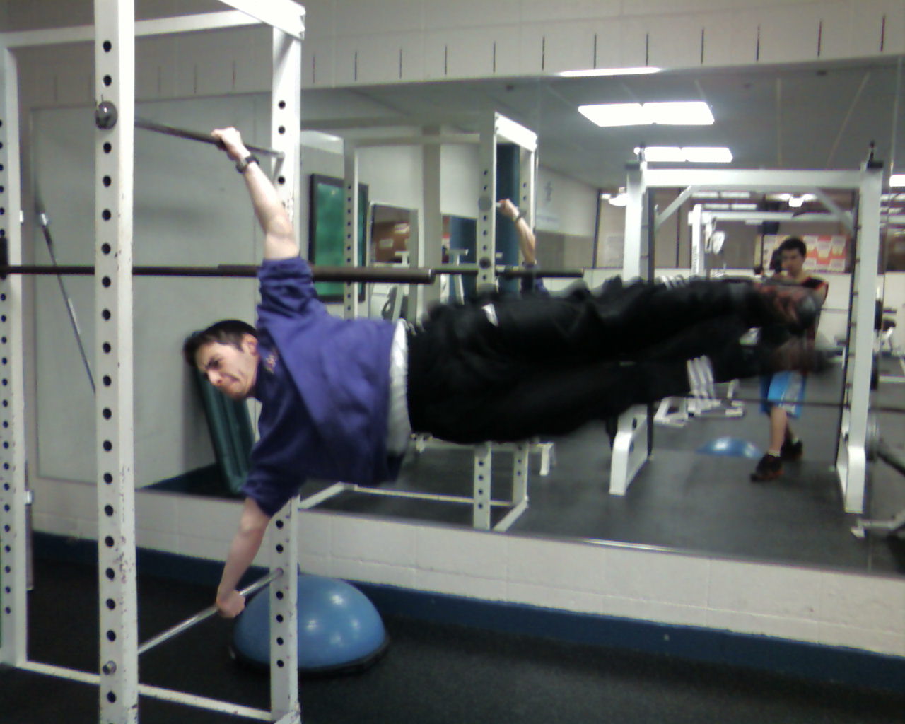Person performing a side plank exercise with one arm on the floor and the other holding a barbell, in a gym.